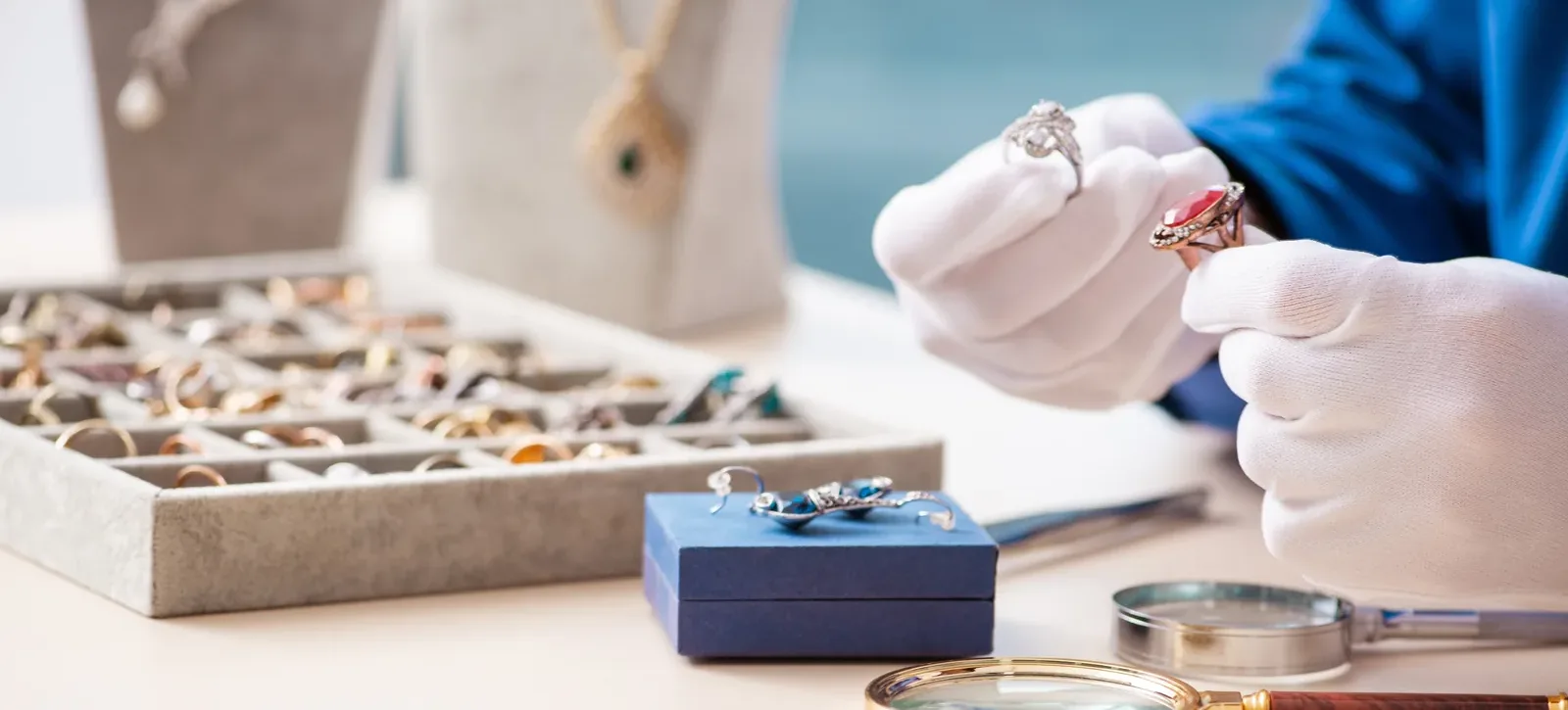 Jeweller in white gloves examining antique diamond and ruby rings, with a magnifying glass and tray of rings on the counter